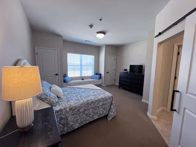 A bedroom with a bed covered in a gray and white patterned bedspread, a bedside table with a lamp, a window with a cushioned bench underneath, two closed doors, a black dresser with a TV on top, and a sliding barn door leading to another room.