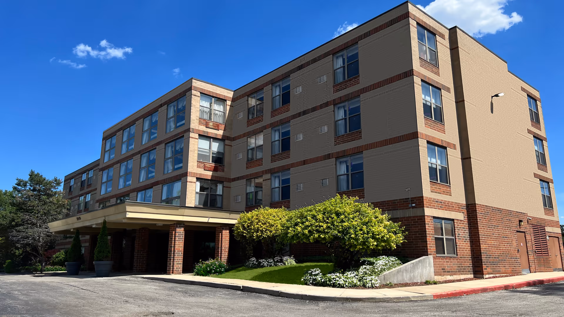 Exterior view of a multi-story senior living facility building with beige and brick facade under a clear blue sky. There are some green shrubs and trees near the entrance with a covered drop-off area.
