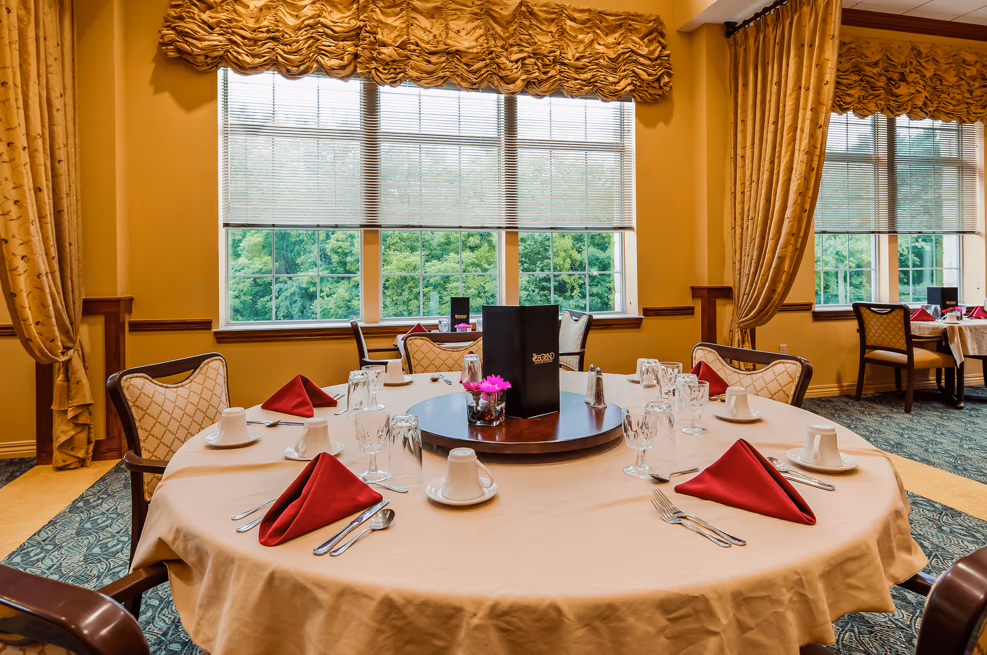 A round dining table set for a meal with beige tablecloth, red folded napkins, white cups and saucers, silverware, and glassware. The table has a centerpiece with a small flower arrangement and a menu labeled 'Legend'. The room features large windows with blinds and gold curtains, and upholstered chairs with a patterned fabric.