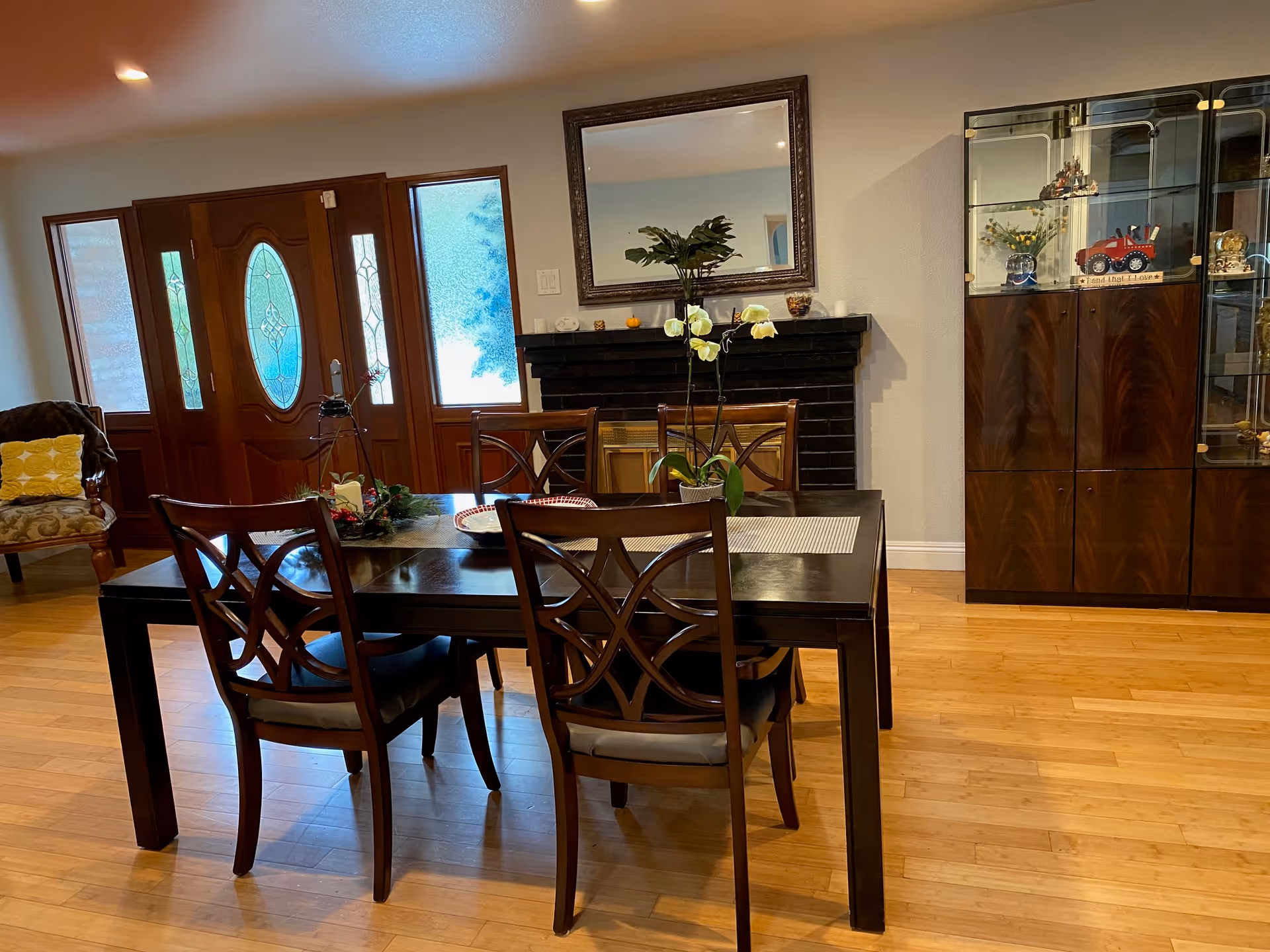 A dining area with a dark wooden table and four matching chairs. The table has a centerpiece with a potted orchid and other decorative items. Behind the table is a black fireplace with a large mirror above it. To the right is a wooden cabinet with glass doors displaying various decorative items. To the left, there is a wooden front door with frosted glass panels and a cushioned chair with a yellow pillow. The floor is light-colored wood.
