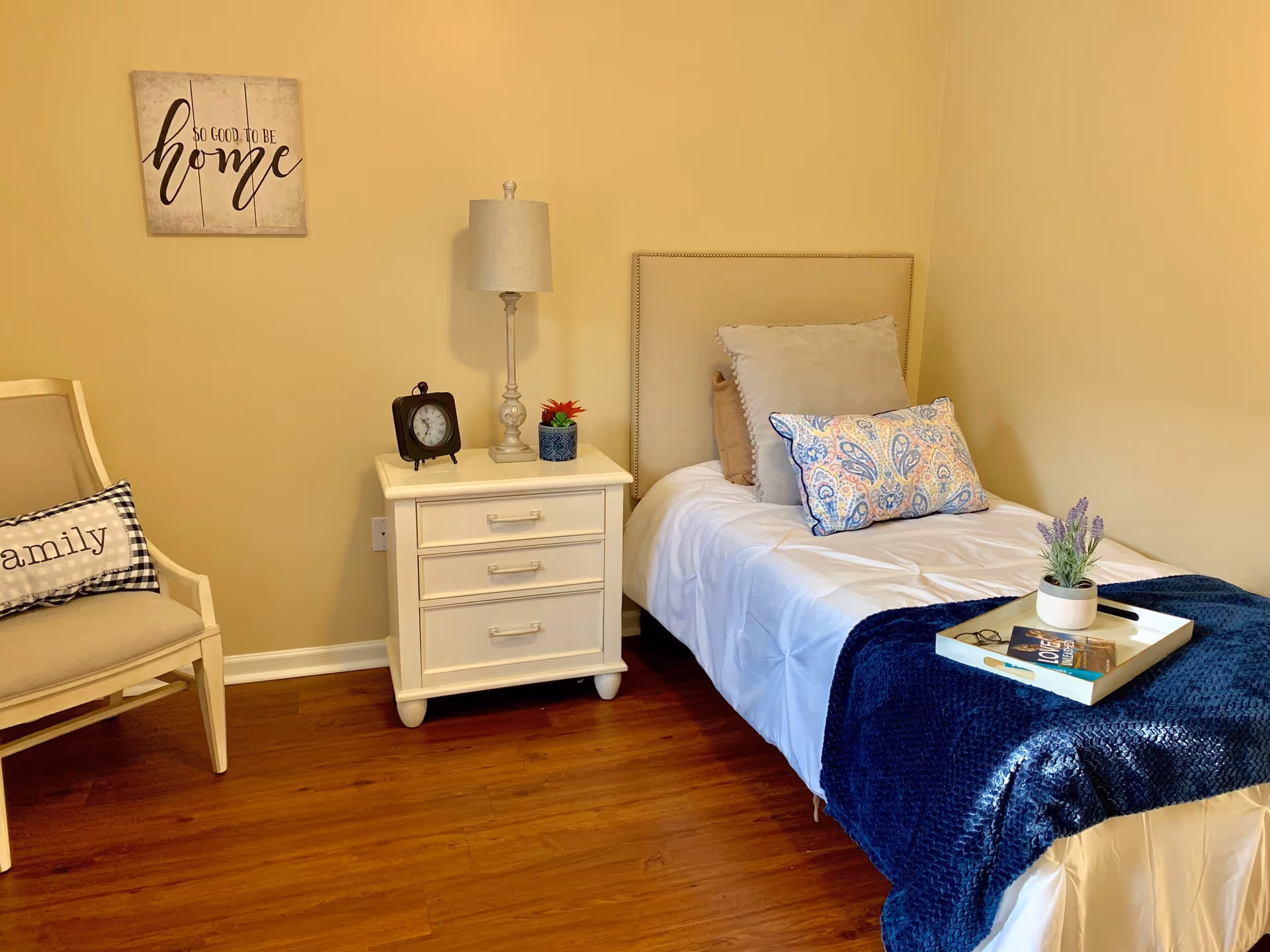 A cozy bedroom with a single bed covered in white bedding and a dark blue throw blanket. The bed has three decorative pillows, and a white tray with a small plant, a book, and glasses rests on the blanket. Next to the bed is a white nightstand with three drawers, holding a table lamp, a small clock, and a small potted plant. To the left, there is a beige armchair with a black and white checkered pillow that says 'family'. A wall decoration above the nightstand reads 'So good to be home'. The room has wooden flooring and light beige walls.