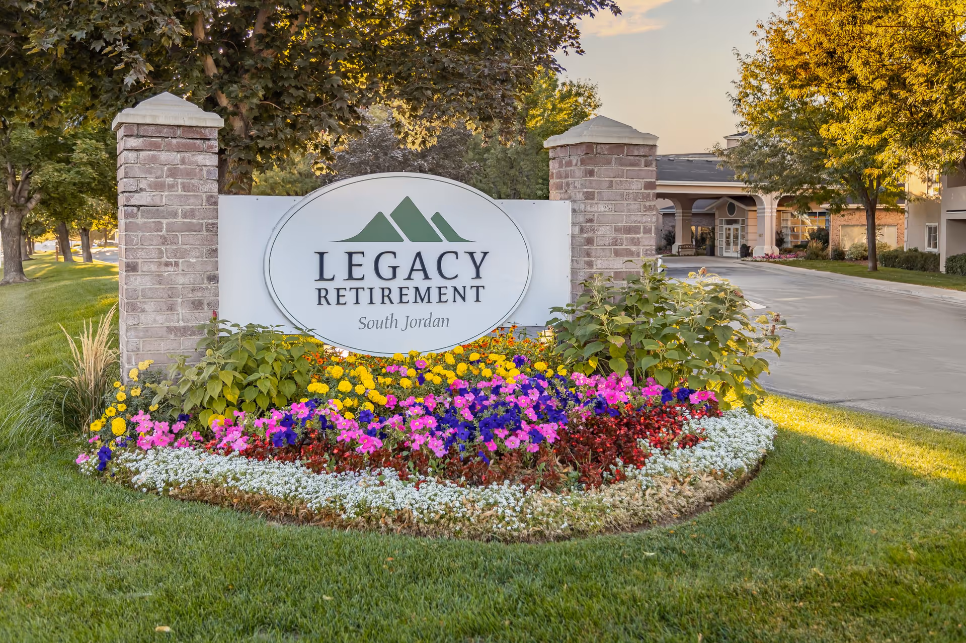 Entrance sign reading "Legacy Retirement South Jordan" set between brick pillars with colorful flowers and the facility building visible behind it.