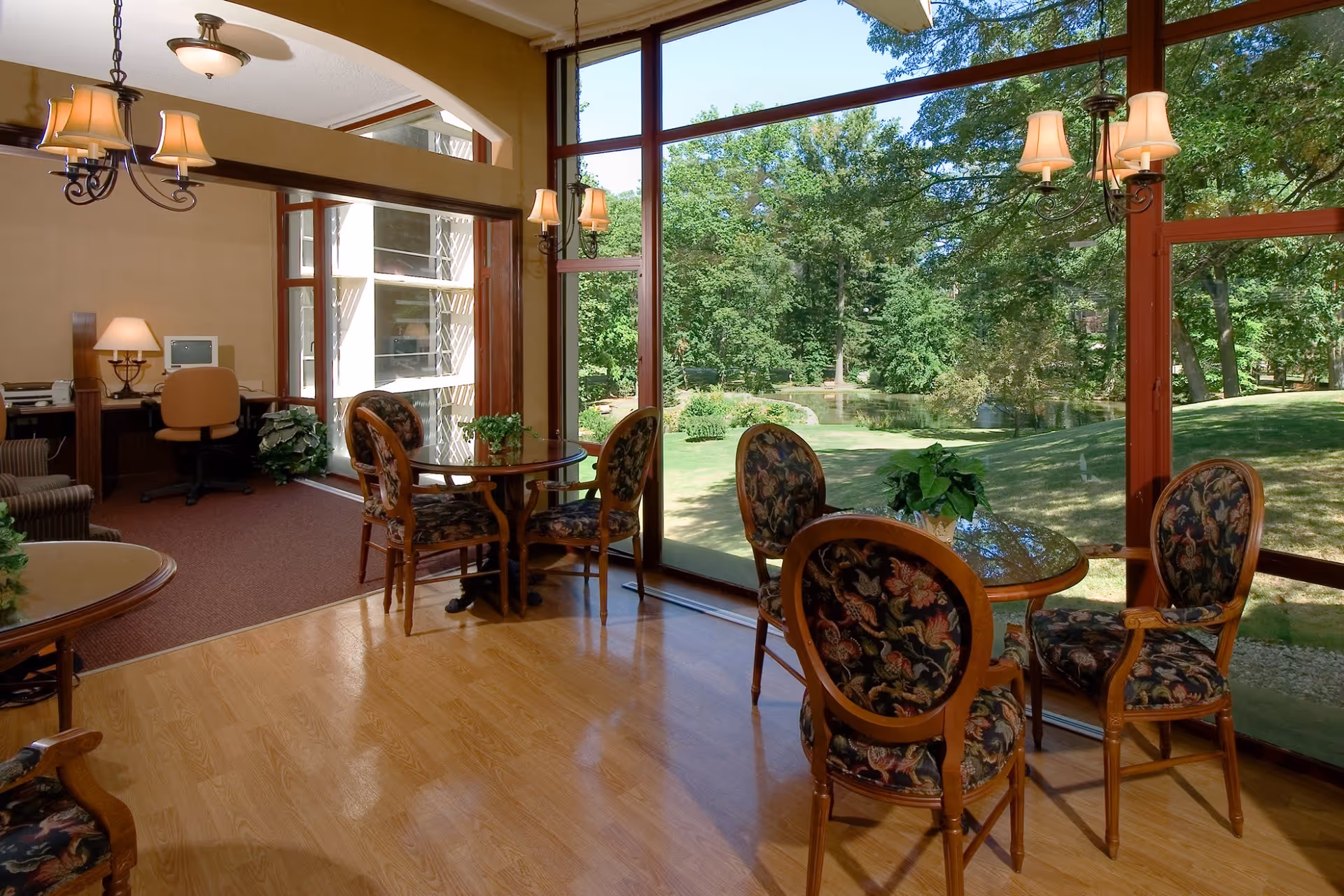 Sunny common room with round tables and upholstered chairs facing large windows overlooking a grassy wooded pond.