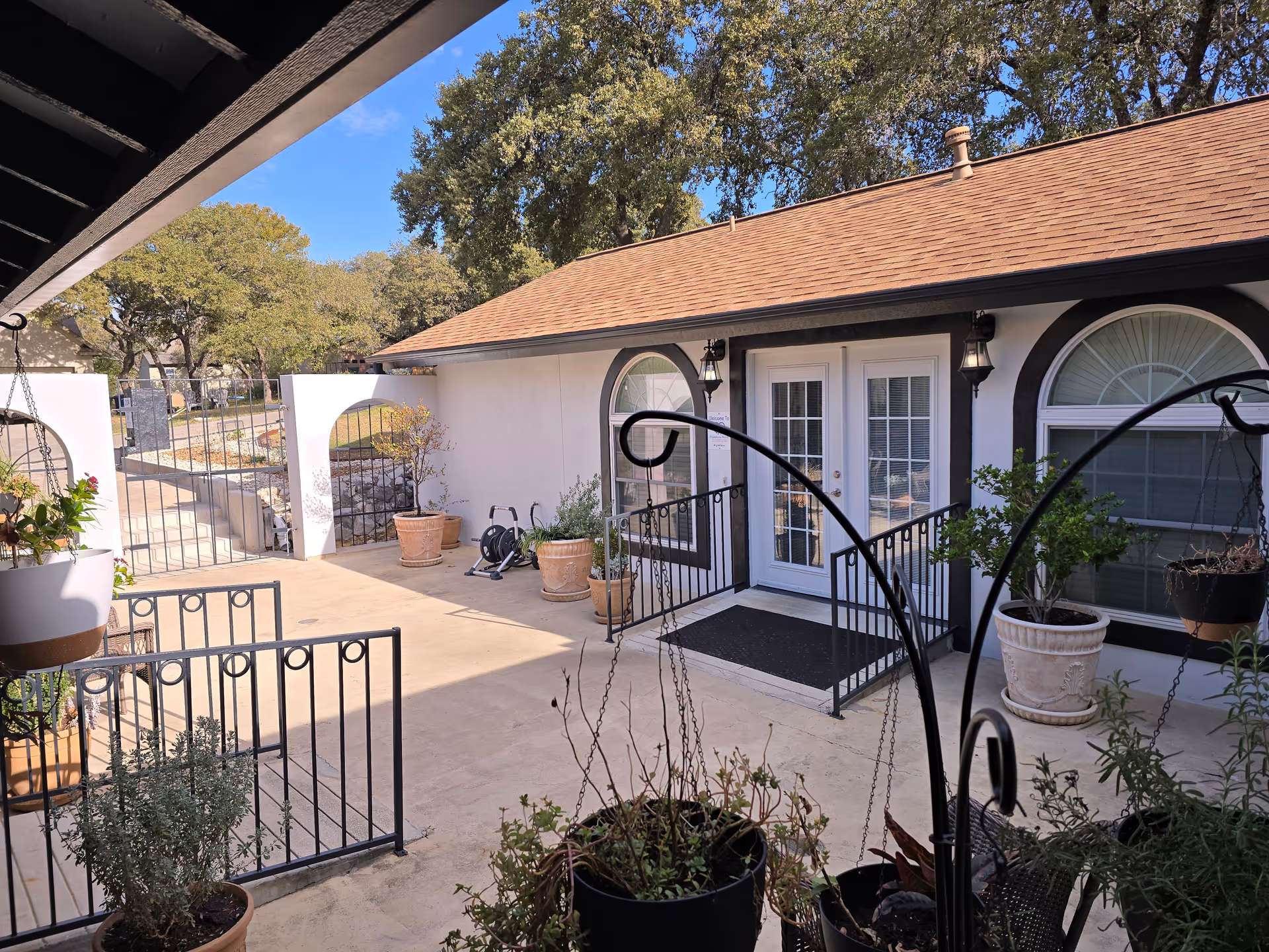 Sunlit courtyard patio with potted plants and a white building featuring double French doors and arched windows.