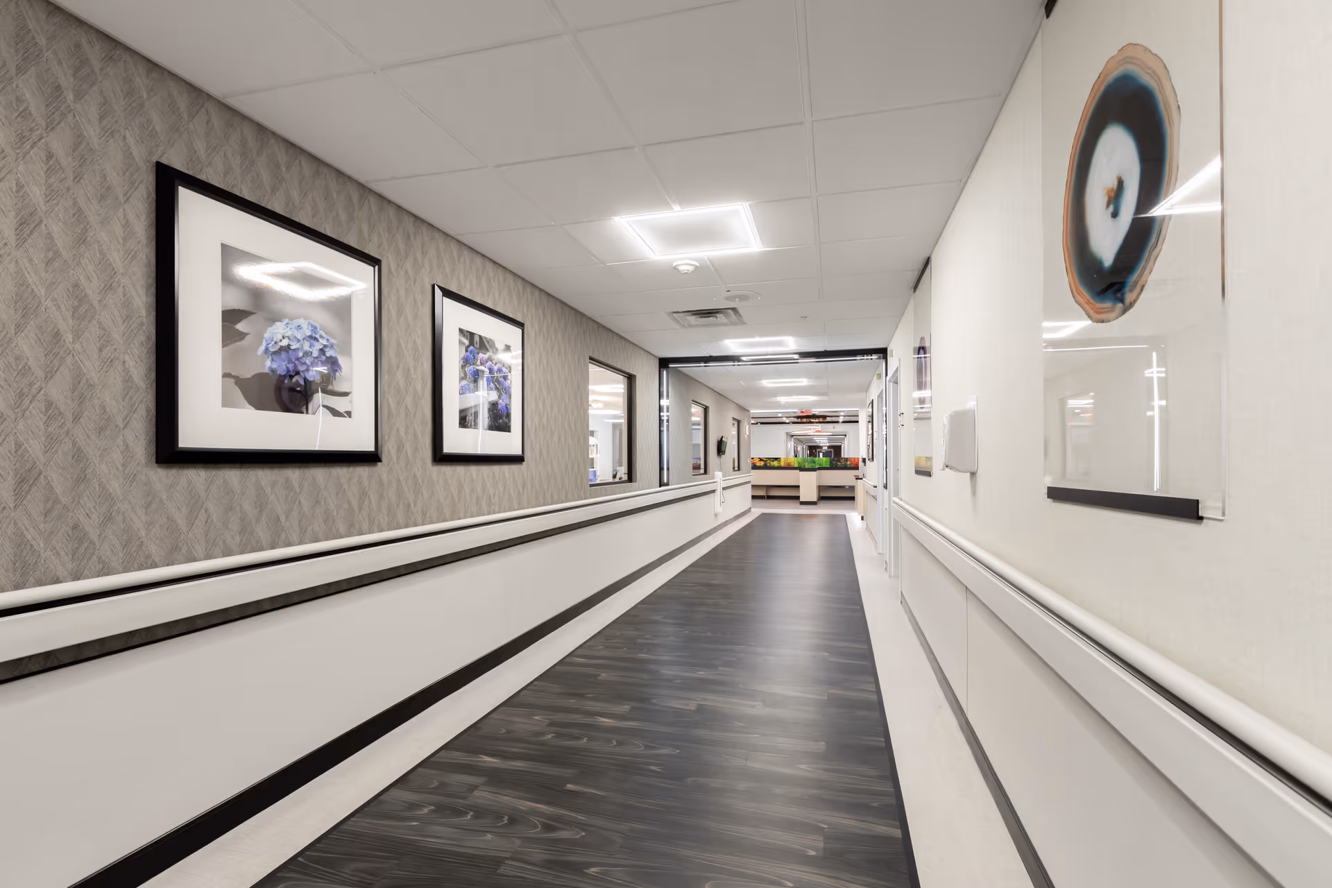 Well-lit long interior hallway in a rehabilitation and nursing facility with framed artwork, handrails, and dark wood-look flooring.