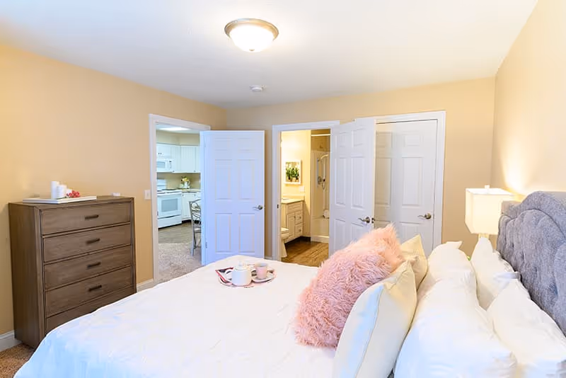A cozy bedroom with a large bed featuring white bedding and pink fluffy pillows. A wooden dresser is on the left side of the room. Two open doors reveal a kitchen area and a bathroom with a shower. The walls are painted beige and the room is softly lit by a ceiling light and a bedside lamp.