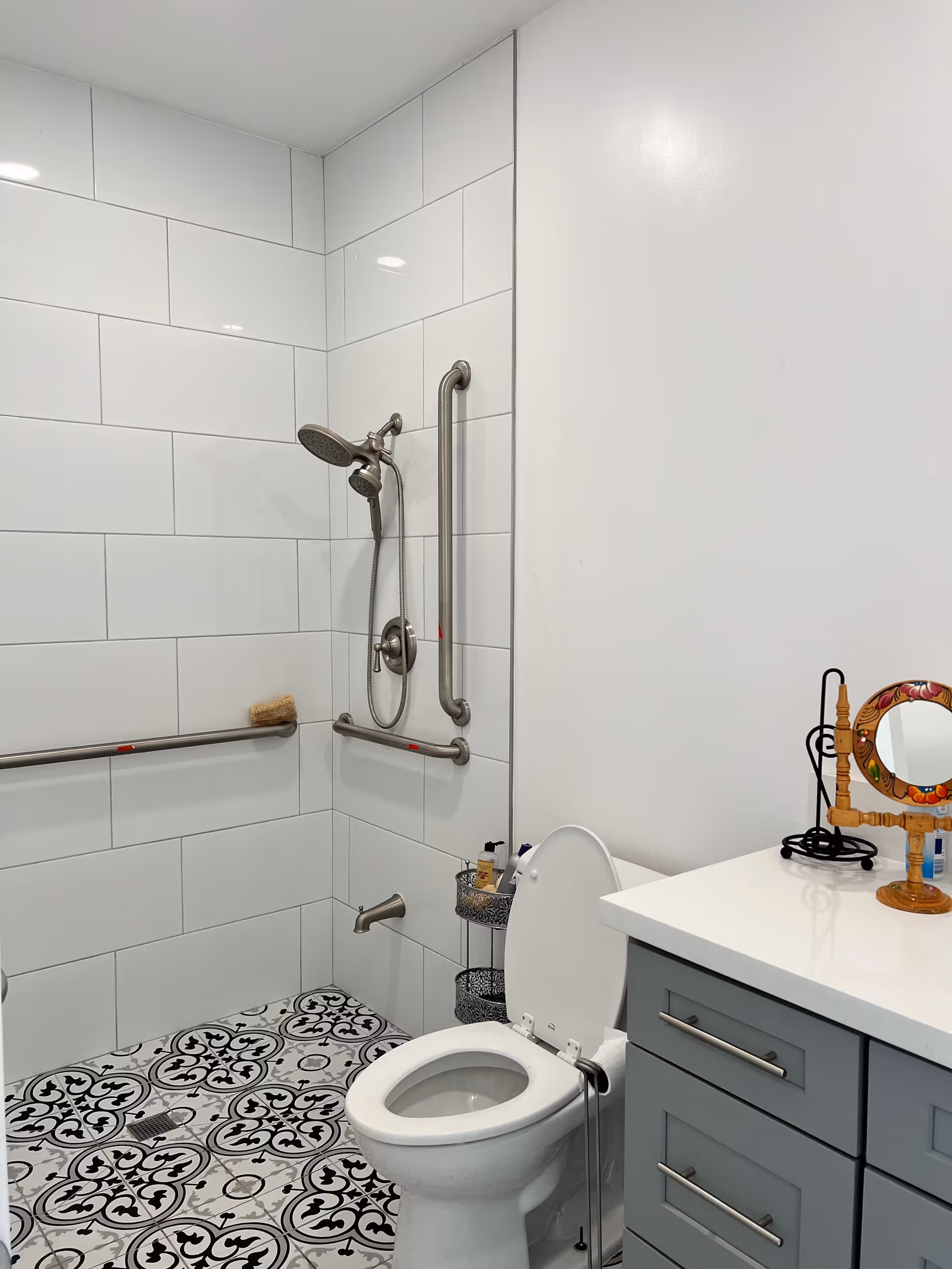 A clean, modern bathroom with white tiled walls and decorative black and white patterned floor tiles. The bathroom features a toilet with the lid open, a shower area with a handheld showerhead and grab bars for accessibility, and a gray vanity with a white countertop. On the countertop, there is a small decorative mirror and a black metal stand.
