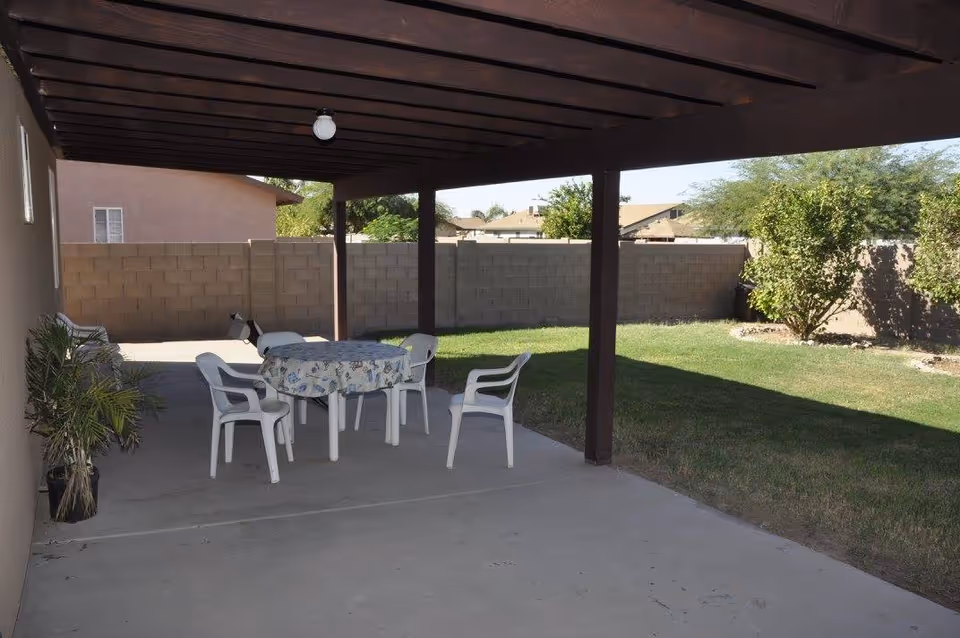 Covered patio with a table and plastic chairs next to a grassy backyard and cinderblock fence.