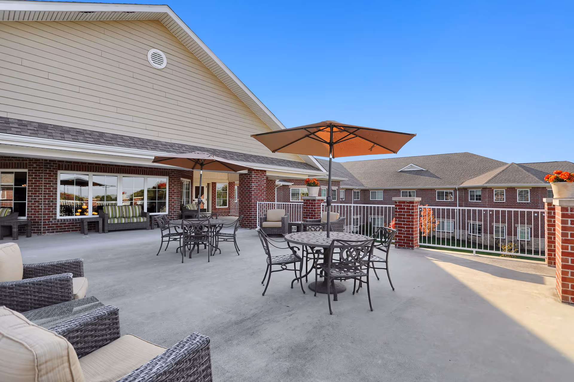 Outdoor patio area at Magnolia Springs Bridgewater with metal tables and chairs, each shaded by large umbrellas. The patio is surrounded by a white railing and brick pillars with flower pots on top. The building exterior features red brick and beige siding under a clear blue sky.