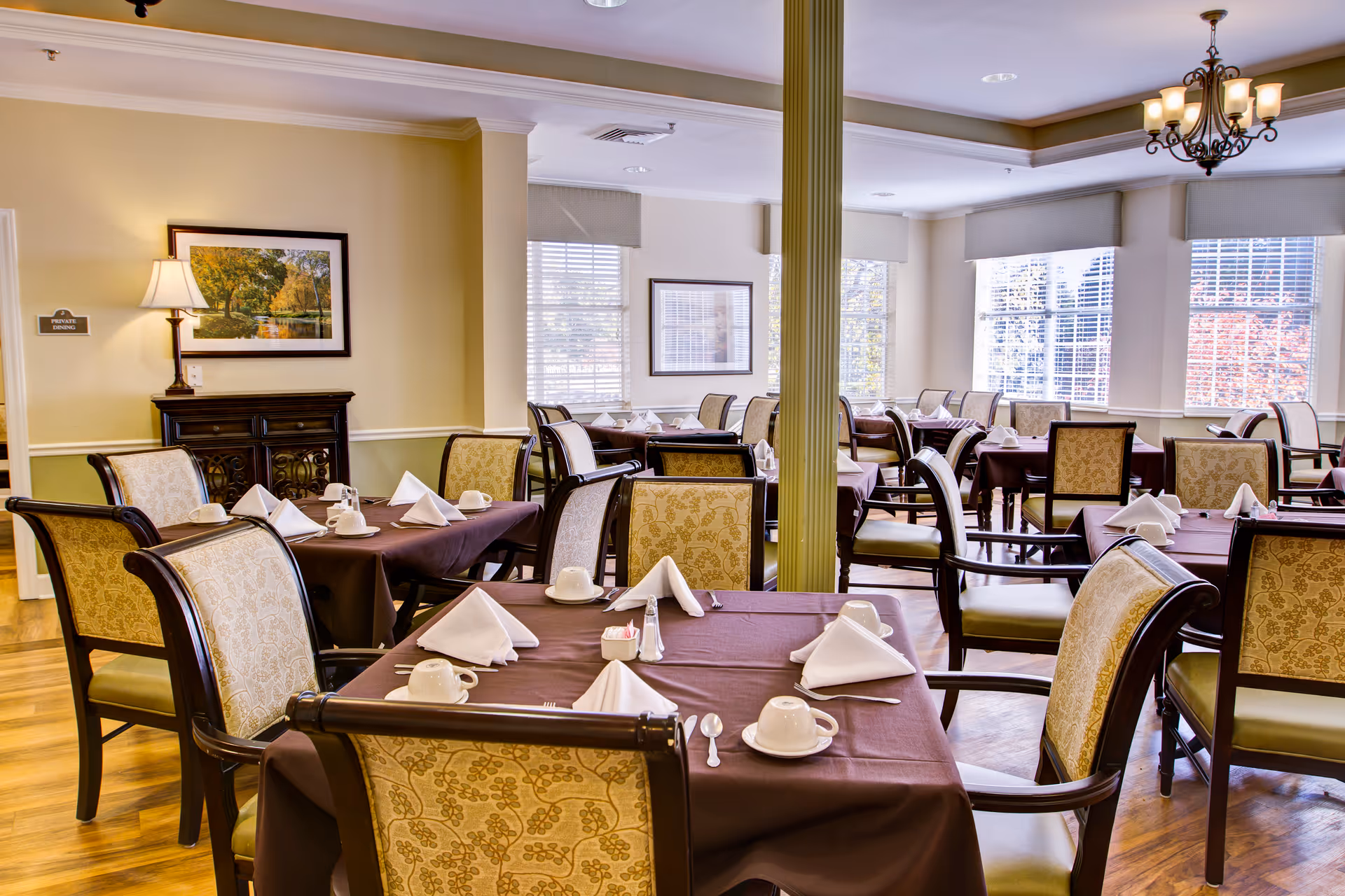 A bright dining room with multiple tables covered in brown tablecloths, each set with white folded napkins, cups, and silverware. The room has large windows with blinds, patterned upholstered chairs, a chandelier, and framed artwork on the walls.