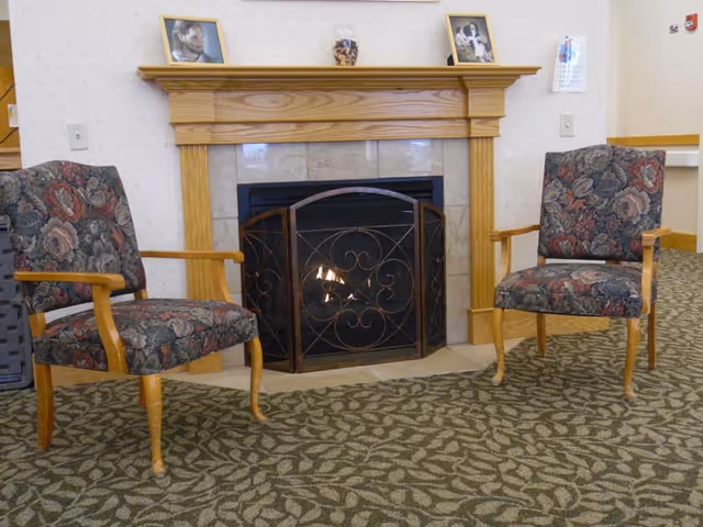 Two upholstered armchairs flank a lit fireplace with a wooden mantel and framed photos in a seating area.
