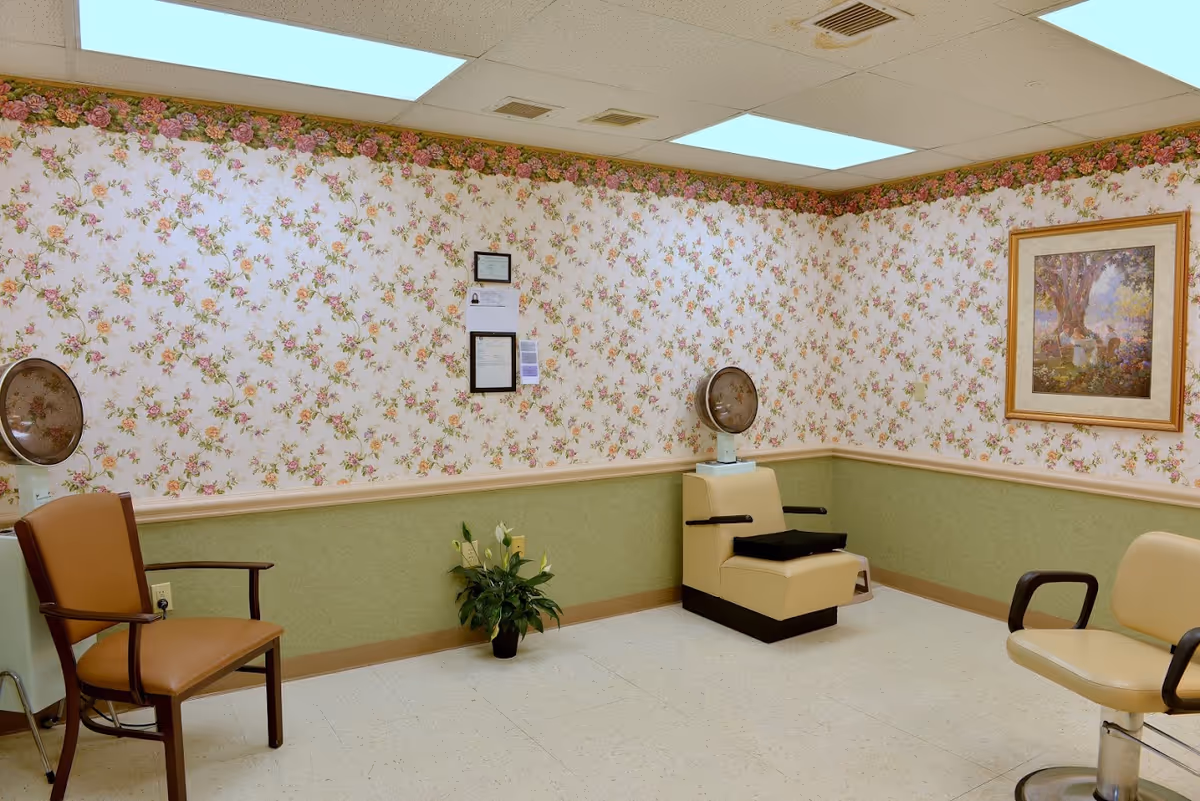 Small interior salon/waiting room with floral wallpaper, salon chairs, hooded hairdryers, and framed artwork.
