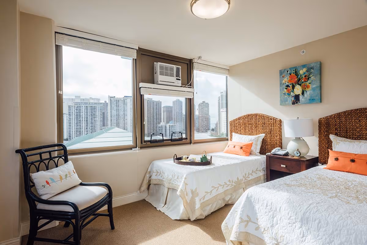 Bright twin-bedded bedroom with wicker headboards, a nightstand, and large windows showing a city skyline.