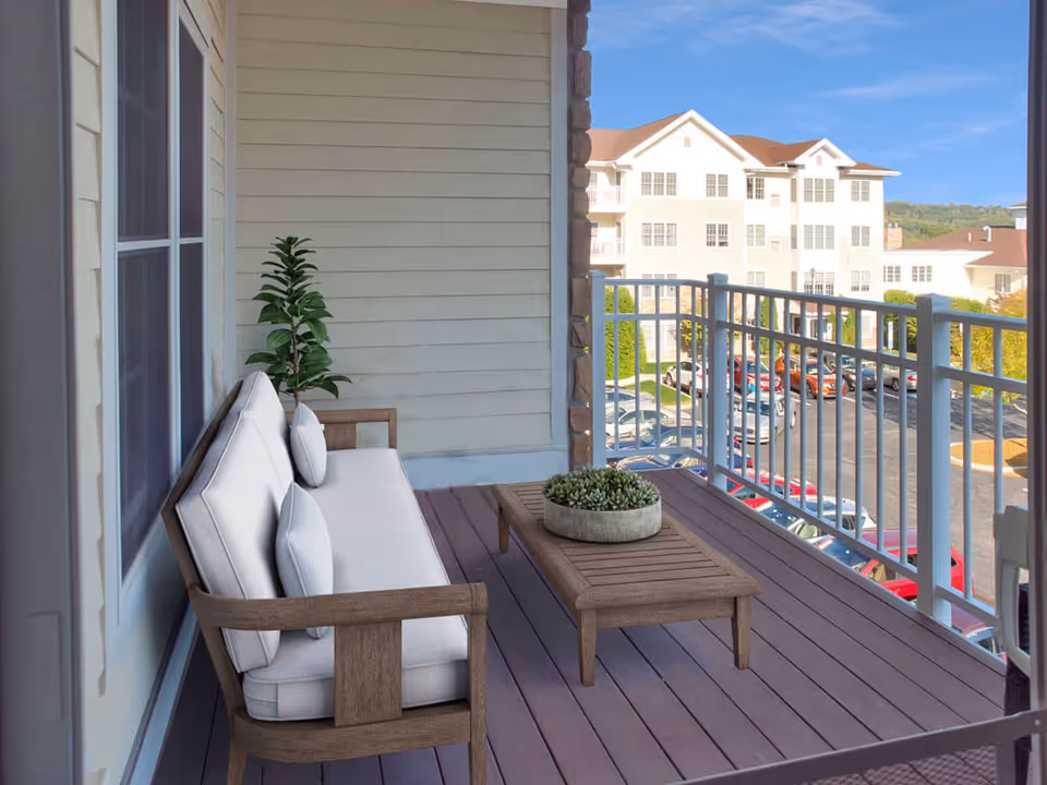 A balcony with a wooden bench that has white cushions and pillows, a wooden coffee table with a round planter containing succulents, a potted plant in the corner, and a view of a parking lot and multi-story residential buildings under a clear blue sky.