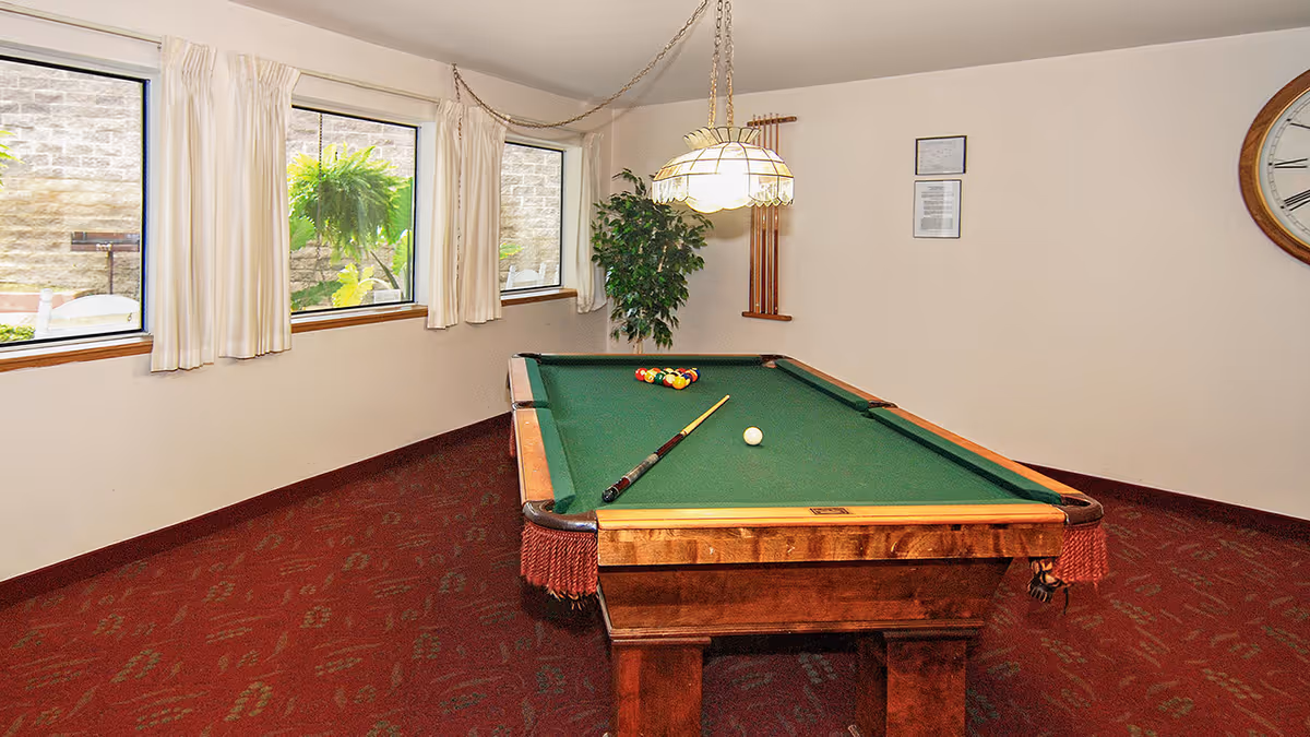 Indoor recreational room with a pool table under a hanging lamp, windows with curtains, and red patterned carpet.