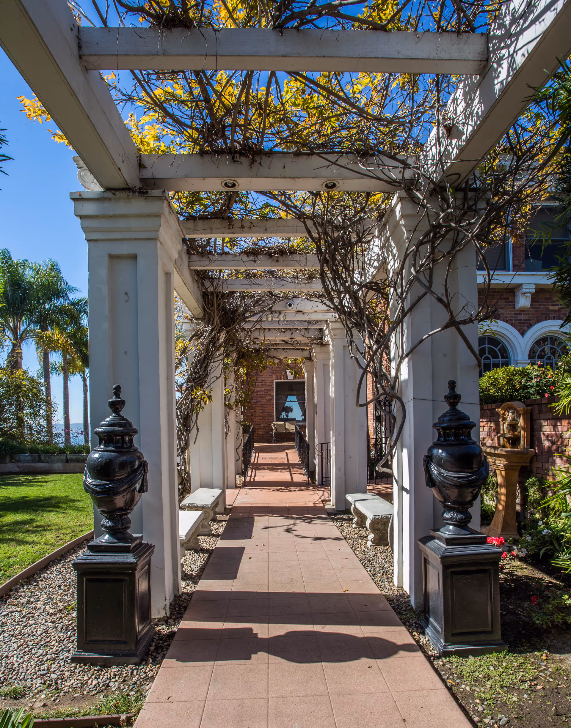 A sunny outdoor walkway with a white pergola covered in climbing plants. The path is paved with tiles and flanked by two black decorative urns on pedestals. There are green lawns, palm trees, and a brick building with arched windows in the background.