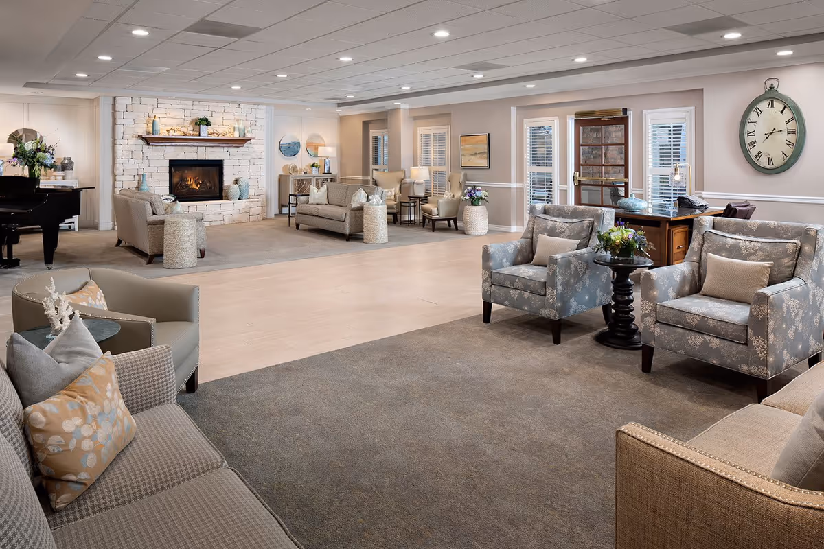 A spacious and well-lit senior living facility common area featuring multiple comfortable armchairs and sofas arranged around a central open space. There is a stone fireplace with a wooden mantel, decorative vases, and plants. The room has a neutral color palette with beige walls, carpeted and wooden flooring, a large wall clock, and a piano in the corner.