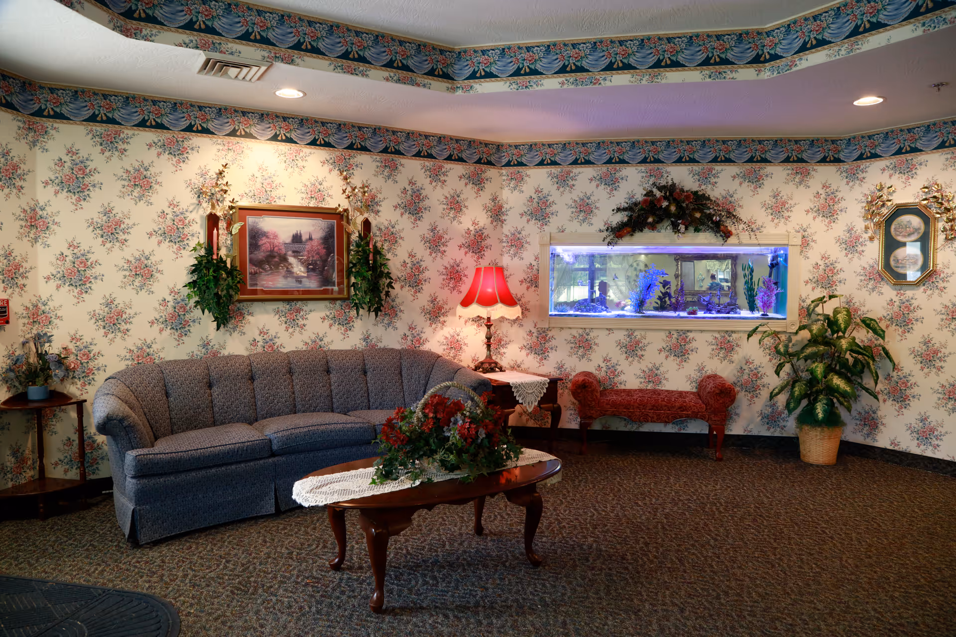 A cozy living room area with floral wallpaper and a patterned carpet. There is a curved gray sofa, a wooden coffee table with a floral centerpiece and lace doily, a side table with a red lamp, and a built-in aquarium with colorful fish. The room also features framed artwork, decorative plants, and a small upholstered bench beneath the aquarium.