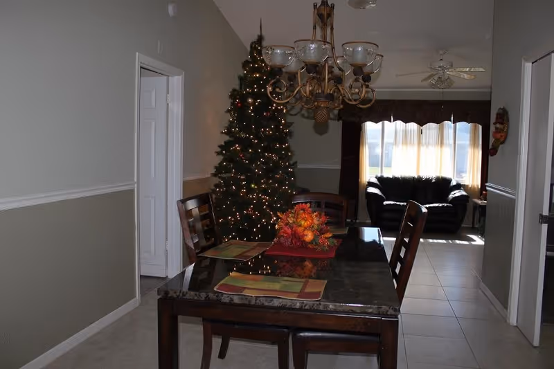 Interior view of a dining area with a dark wooden table set with placemats and a fall-themed centerpiece. To the left, there is a decorated Christmas tree with lights. In the background, a living room area is visible with a black leather couch and large windows with sheer curtains allowing sunlight to enter. A chandelier hangs above the dining table.