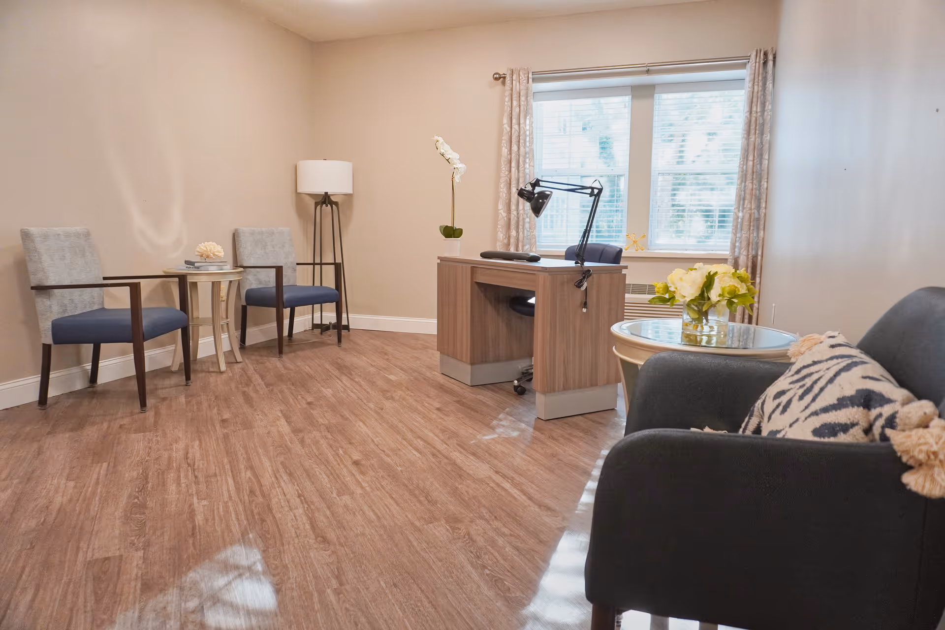 Bright furnished sitting room/office with chairs, a desk, side tables, lamp, and a window in an assisted living facility.