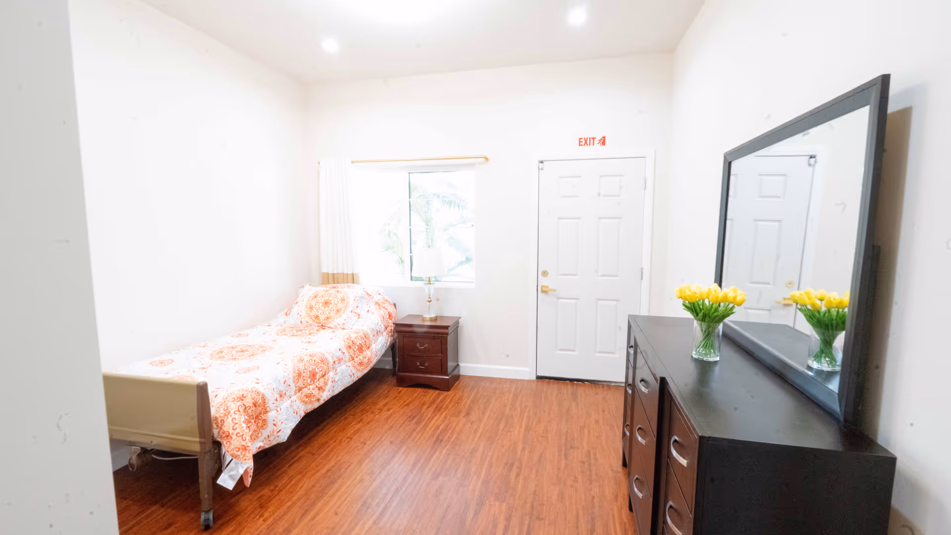 A simple bedroom with a single bed covered in an orange and white patterned bedspread, a wooden nightstand with a lamp next to the bed, a window with white curtains, a white door with an exit sign above it, and a dark wooden dresser with a large mirror and a vase of yellow flowers on top.