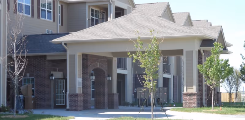 Exterior view of a residential building with a covered entrance supported by columns. The building has brick and siding walls, multiple windows, and a sloped roof. There are young trees and a mailbox cluster in front of the entrance.