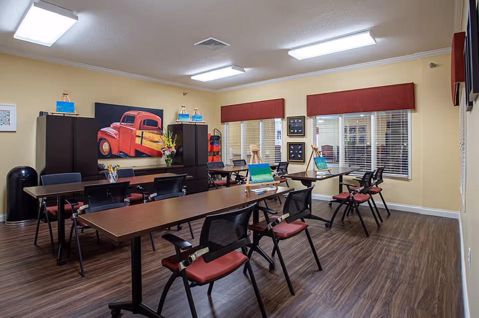 A well-lit activity room with several tables and chairs arranged for painting. Each table has small easels with canvas paintings in progress. The room has wood flooring, beige walls, and windows with red valances. A large painting of a red vintage truck is displayed on the back wall above black cabinets.