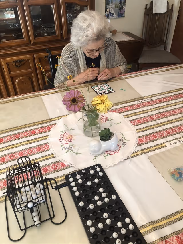 An elderly woman with white hair and glasses is sitting at a table playing bingo. The table is covered with a patterned tablecloth and has a vase with flowers and a small potted plant on a doily. A bingo cage with balls and a bingo board with markers are also on the table. Behind her is a wooden cabinet and a chair.