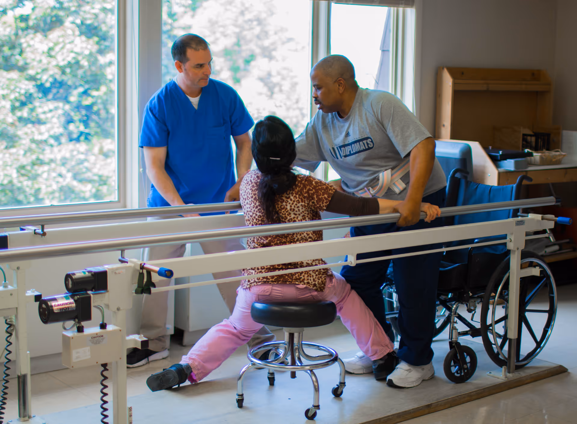 A woman is performing physical therapy exercises using parallel bars with assistance from two men, one in blue scrubs and the other in casual clothing, in a bright room with large windows and a wheelchair nearby.