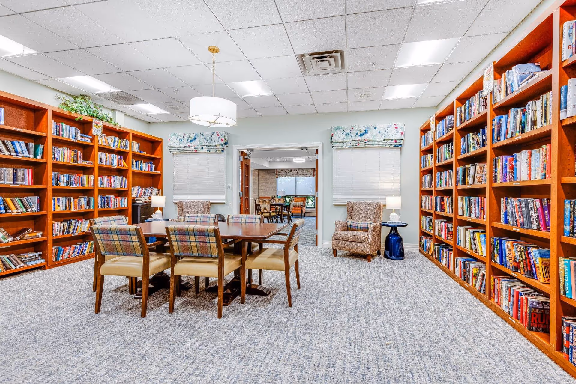 A bright and spacious library room with wooden bookshelves filled with books lining both walls. In the center is a wooden table surrounded by six plaid upholstered chairs. Two armchairs with plaid cushions are placed near windows with floral valances. A modern white pendant light hangs from the ceiling, and a blue side table with a lamp is next to one armchair. Through a doorway, another room with additional seating and large windows is visible.