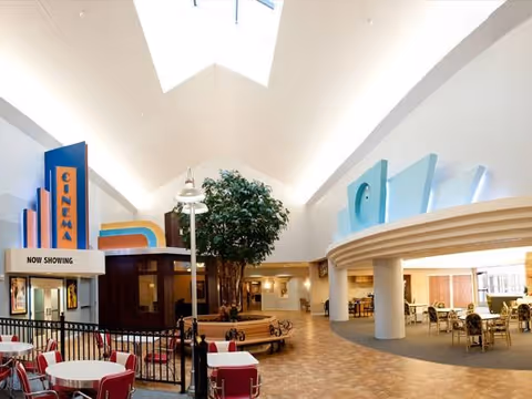Interior view of a senior living facility featuring a small cinema area with a 'Now Showing' sign, round tables with red chairs, a large indoor tree with circular wooden bench seating around it, and an open common area with additional seating and tables under a high ceiling with skylights.