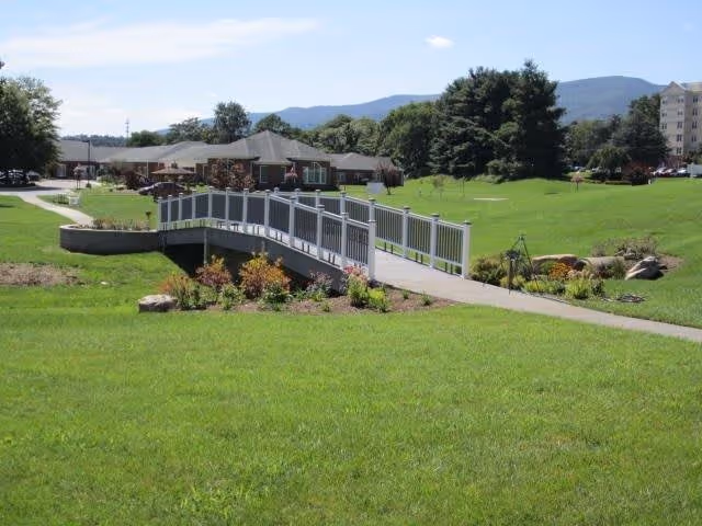 A green outdoor area with a small pedestrian bridge over a landscaped garden bed. In the background, there are buildings, trees, and hills under a clear blue sky.
