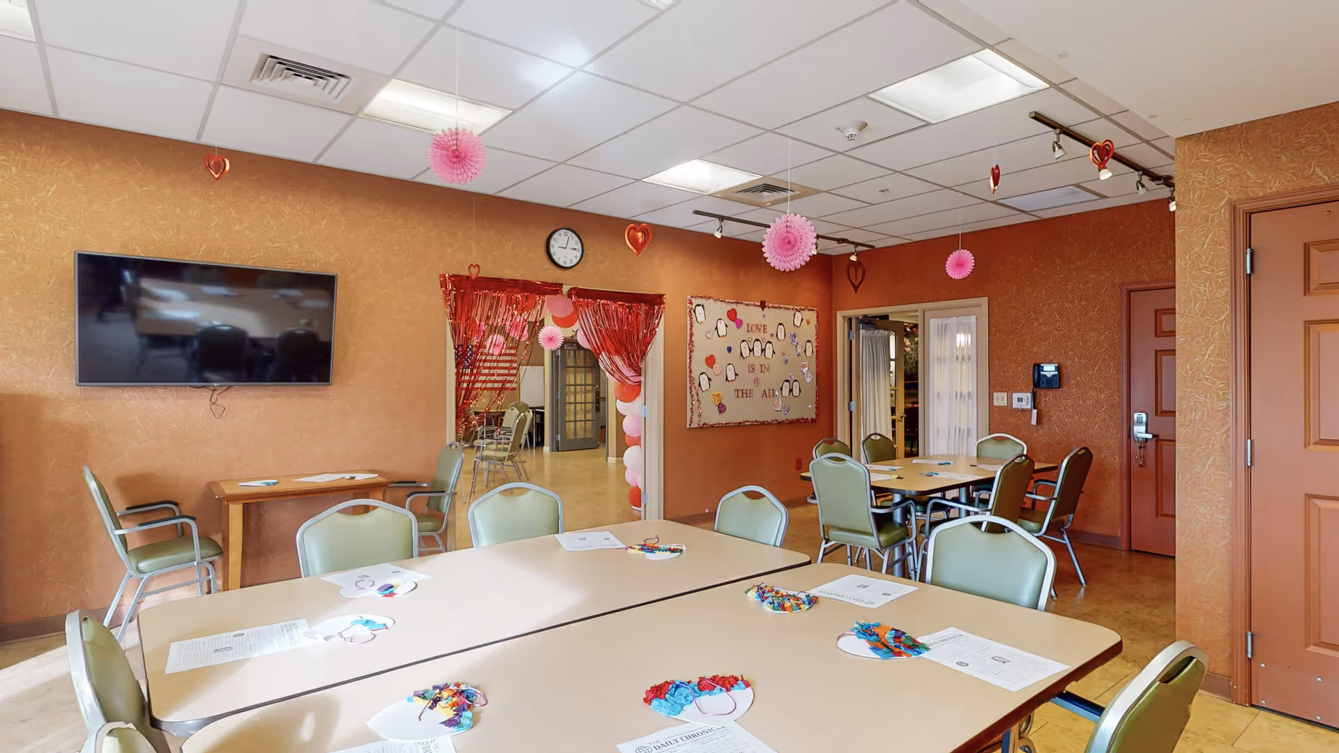 A community room with several tables and green chairs arranged for group activities. The walls are decorated with Valentine-themed decorations including hanging pink and red hearts and paper pom-poms. A bulletin board on the wall reads 'Love is in the air' with heart decorations. A flat-screen TV is mounted on one wall, and there is a clock above a doorway draped with red curtains and balloons. The room has a warm, inviting atmosphere with brownish walls and a tiled floor.