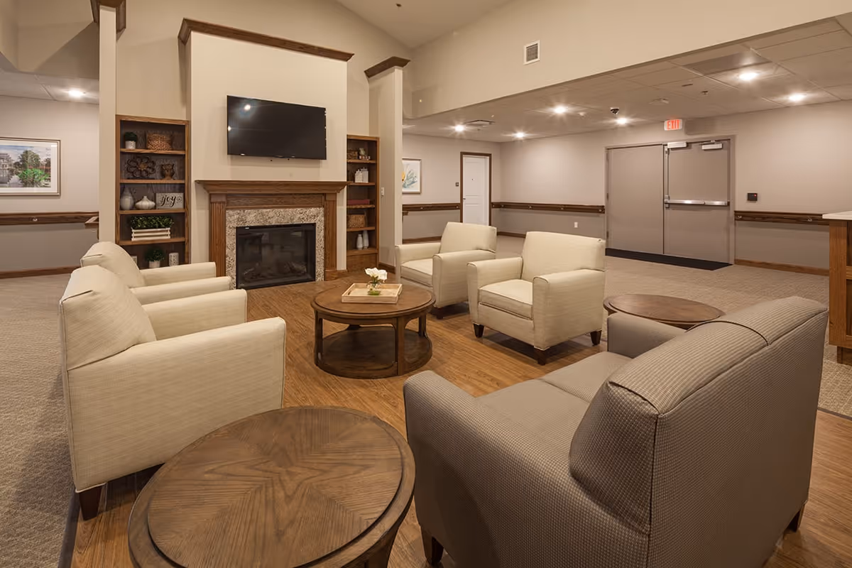 A cozy senior living facility common area with beige armchairs and a sofa arranged around a wooden coffee table. There is a fireplace with a mounted flat-screen TV above it, flanked by built-in shelves with decorative items. The room has warm lighting, wood flooring in the seating area, and carpet in the surrounding space. Double doors and framed artwork are visible in the background.