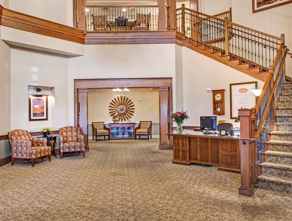 Spacious interior lobby area with a wooden reception desk on the right, a staircase with wooden railings leading to an upper floor, two patterned armchairs with a small table and flowers on the left, and a decorative wall piece with two chairs and a small table in the background.