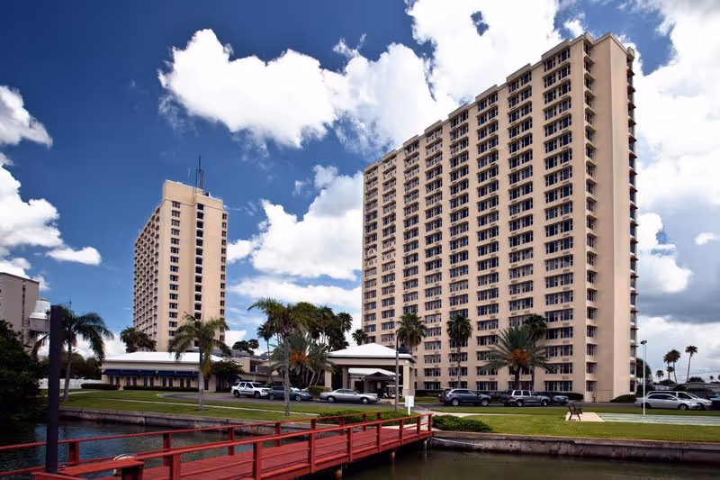 Two tall beige apartment buildings by water with a red bridge and palm trees.