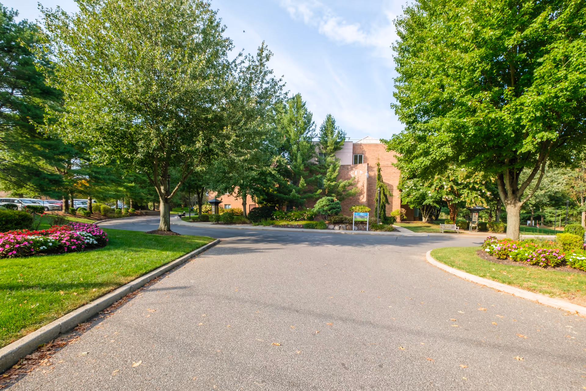 Driveway and landscaped entrance leading to a brick senior living building surrounded by trees and flower beds.
