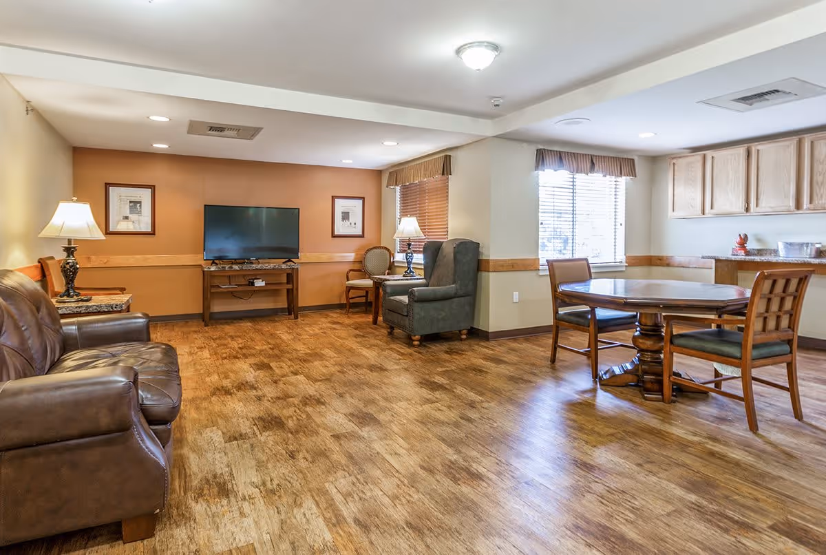A spacious living room area with wooden flooring, featuring a brown leather sofa, an armchair, a round wooden table with four chairs, a TV on a wooden stand, two framed pictures on the wall, and two table lamps. There are windows with blinds and wooden cabinets on the right side.