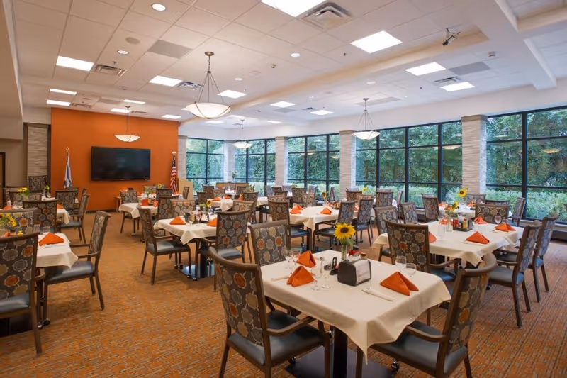 A spacious dining room with multiple tables covered in white tablecloths, each set with orange folded napkins, glassware, and floral centerpieces. The room features large windows along one wall, allowing natural light to illuminate the space. There are patterned chairs around each table, and the walls are painted in warm tones with a mounted flat-screen TV and flags in the background.
