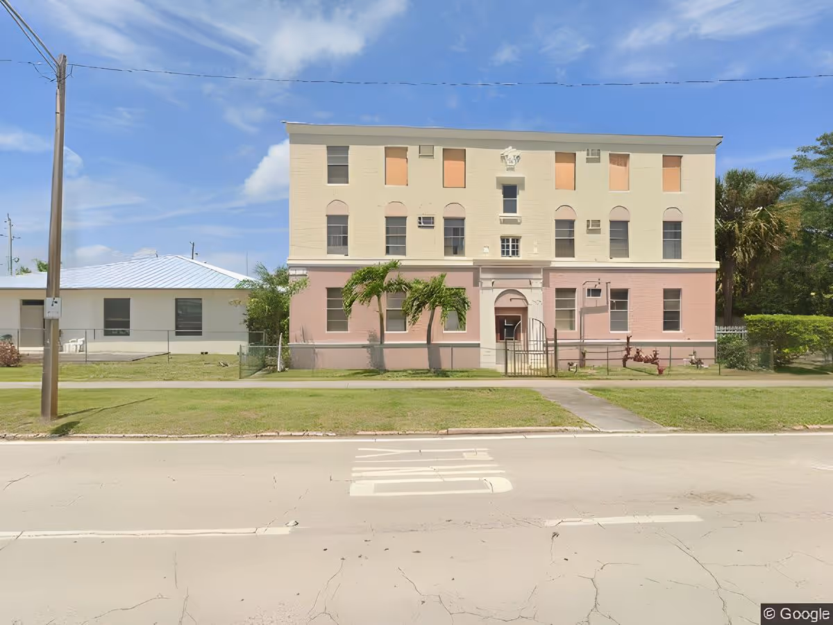 Front view of a three-story cream-and-pink building with a lawn, sidewalk and road in the foreground under a blue sky.