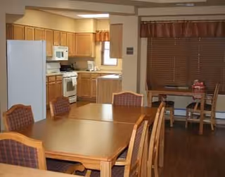 Interior view of a senior community dining area and kitchen. The foreground shows wooden tables and chairs arranged for dining. In the background, there is a kitchen with wooden cabinets, a white refrigerator, microwave, stove, and a small window with a valance. Another table with chairs is positioned near a large window with closed blinds and a valance.