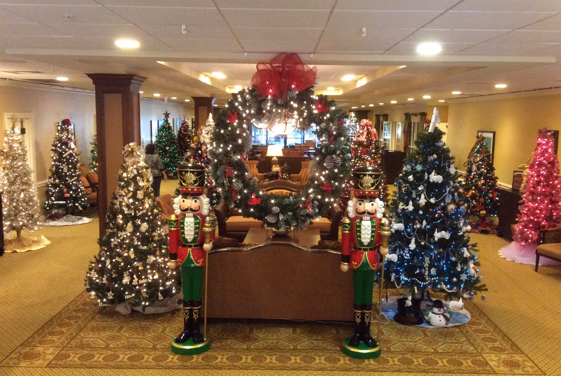 A senior living facility lounge decorated for Christmas with multiple lit trees, a large wreath and two nutcracker statues.