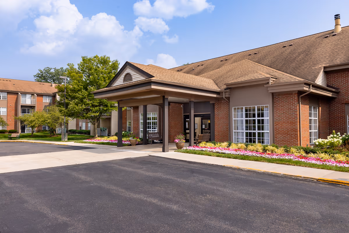 Entrance facade of a brick senior living building with a covered porte-cochère, landscaped flower beds, and a paved driveway.