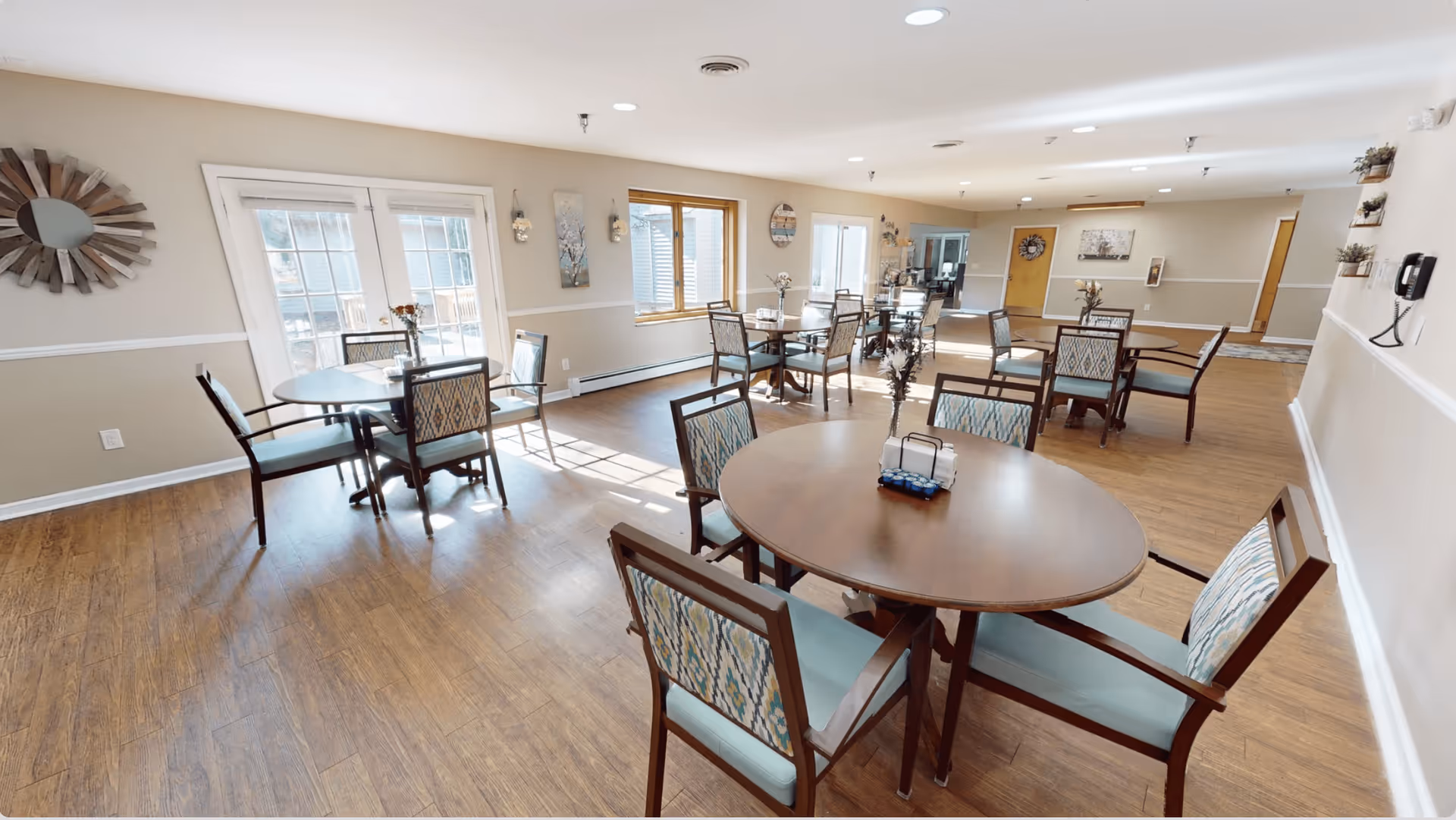 A bright and spacious dining room with multiple round wooden tables and cushioned chairs arranged neatly. The room has large windows and glass doors allowing natural light to fill the space. The walls are decorated with artwork and a decorative mirror, and the floor is wooden.