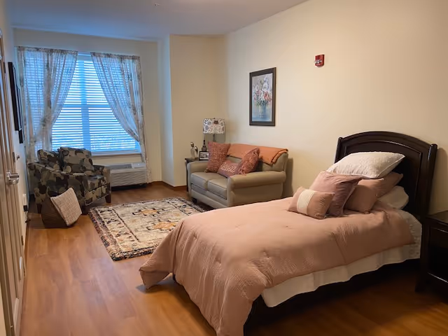 Sunlit studio-style bedroom with a single bed, loveseat, patterned armchair, rug, and a large window with sheer curtains.