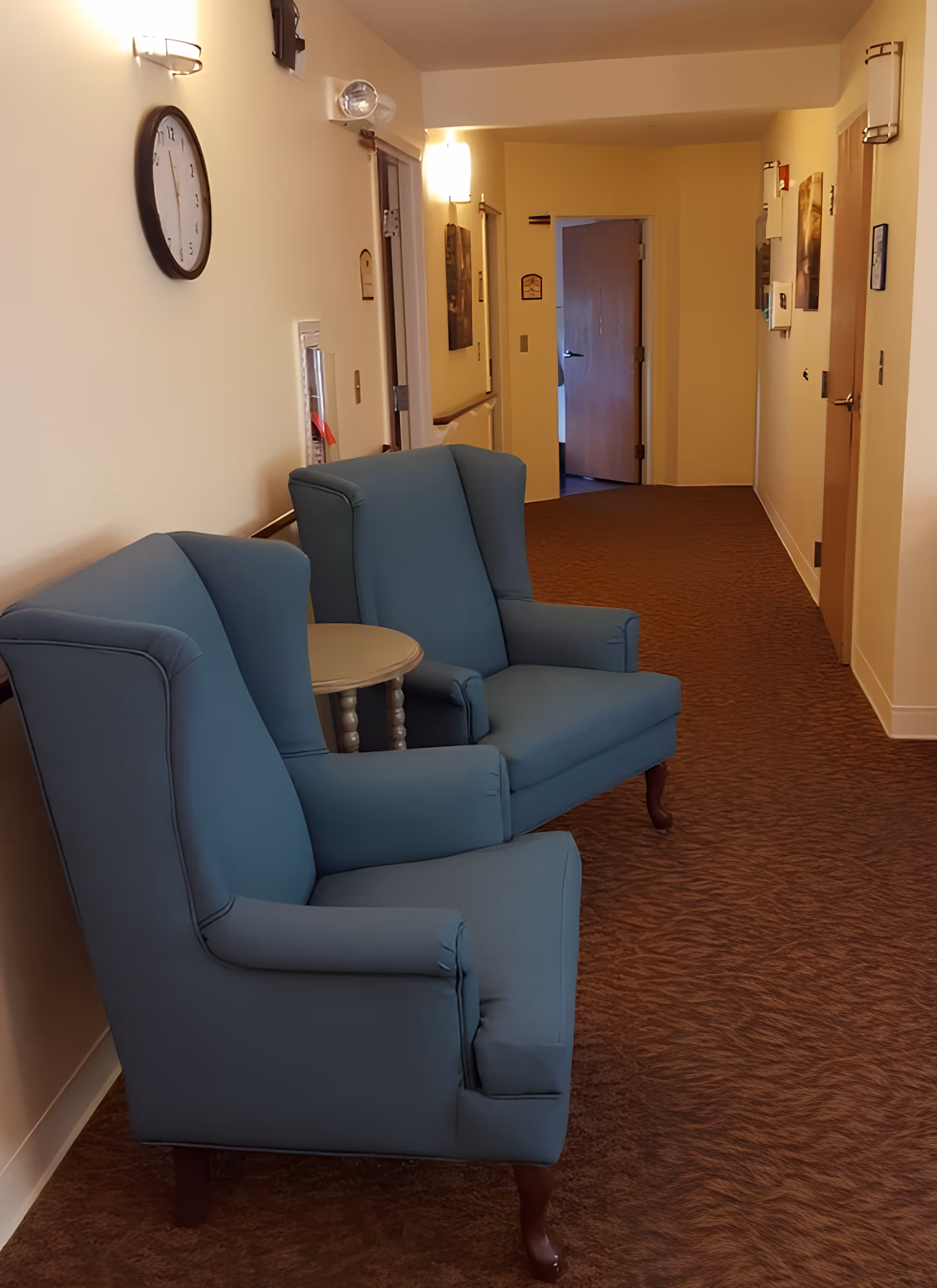 A hallway in a senior living facility with two blue upholstered armchairs and a small round table between them. The walls are light-colored with a clock mounted on the left wall and several light fixtures. The hallway has brown carpet and several doors along the right side, with one door open at the end of the hallway.