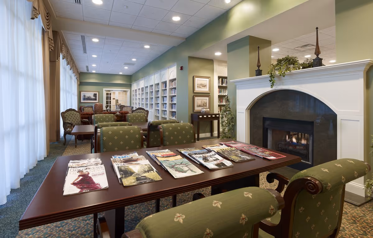Sunlit reading/lounge area with magazines on a table, upholstered chairs, bookshelves and a fireplace.
