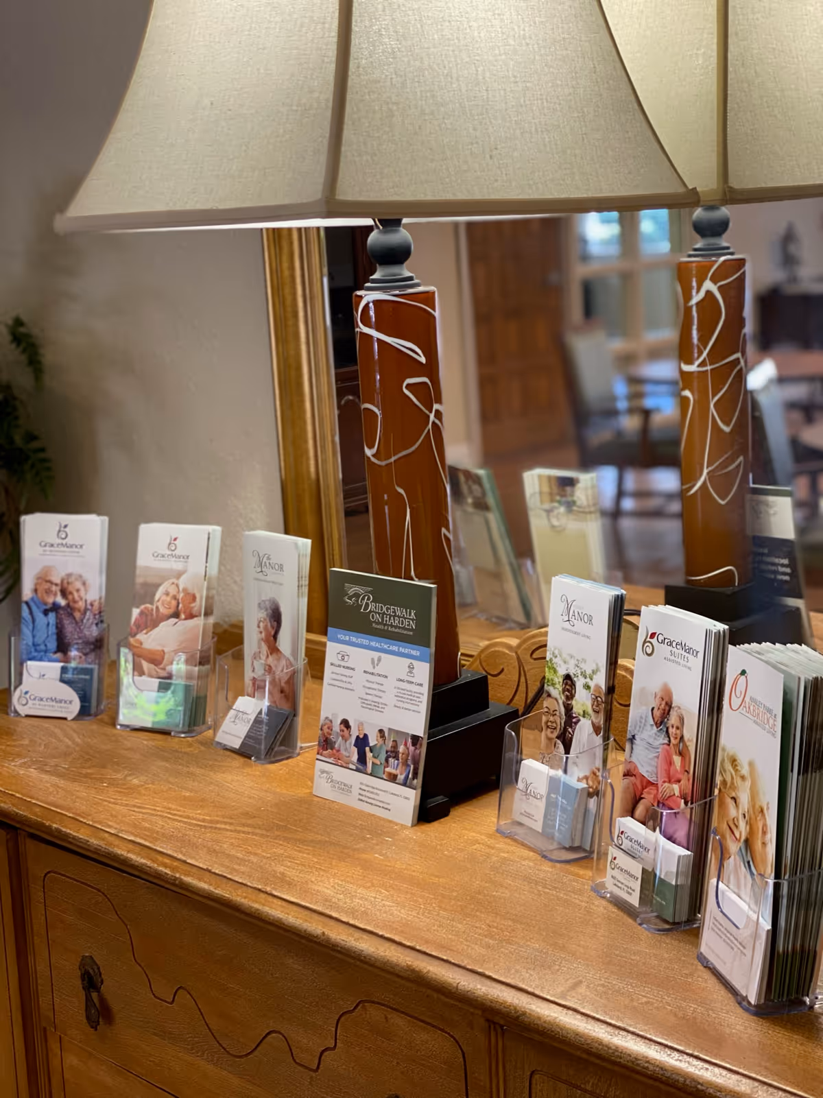 A wooden dresser with a large mirror and two decorative lamps with beige shades. On the dresser are multiple brochure holders containing brochures for senior living facilities, including one prominently labeled Bridgewalk on Harden. The background shows a blurred interior space with chairs and a door.