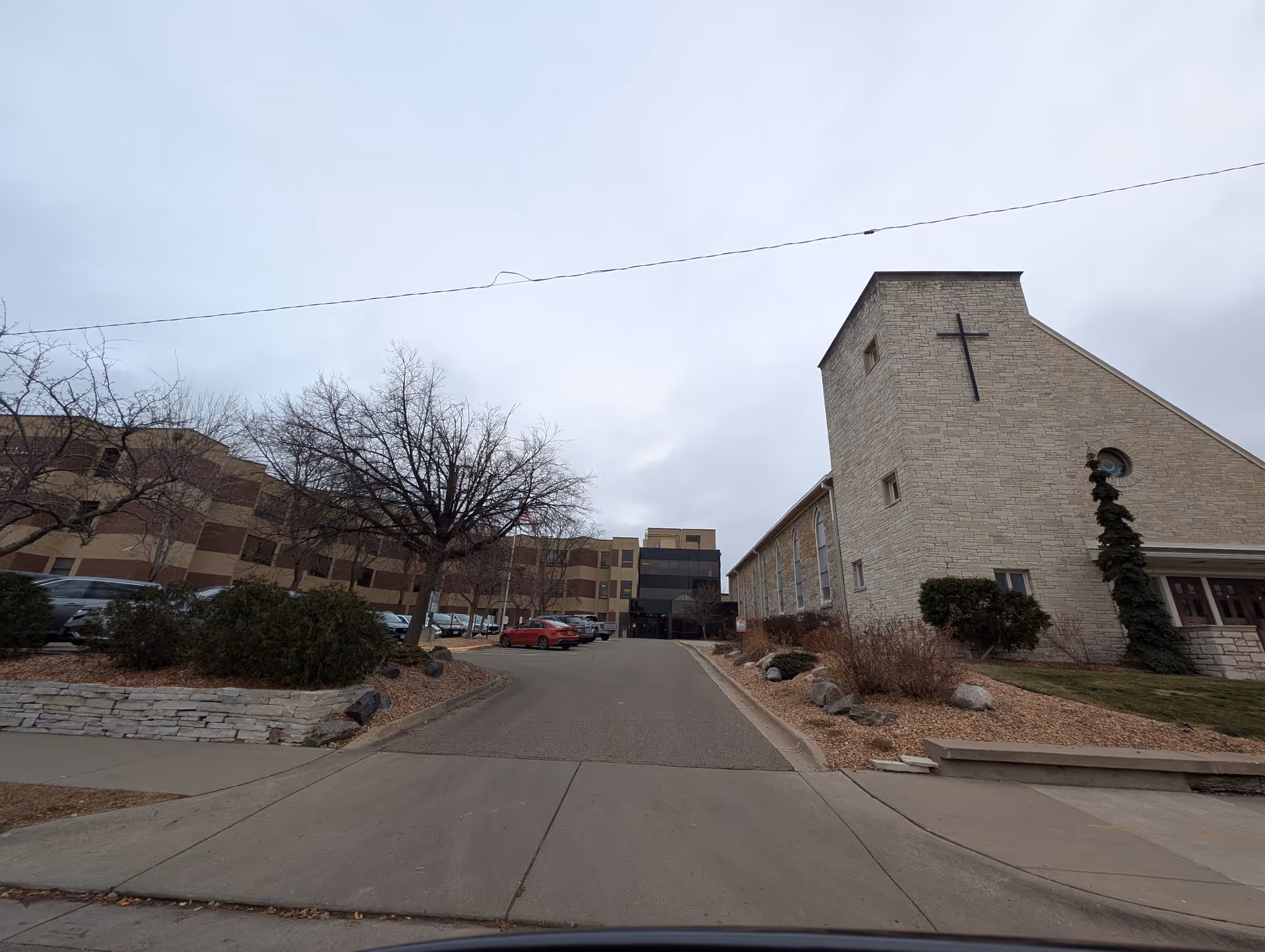Exterior view of a senior living facility with a large multi-story building on the left and a stone building with a cross on the right, under a cloudy sky. There are leafless trees, bushes, and parked cars along the driveway leading to the entrance.