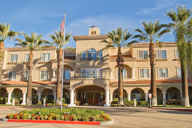 Front exterior view of Bayshire San Dimas facility with a beige multi-story building, palm trees, an American flag, and a landscaped roundabout with flowers under a clear blue sky.